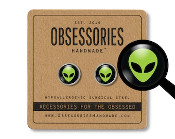 May include: A pair of hypoallergenic surgical steel earrings featuring a green alien head design. The earrings are packaged on a brown card with the text "OBSESSORIES HANDMADE" and "ACCESSORIES FOR THE OBSESSED".