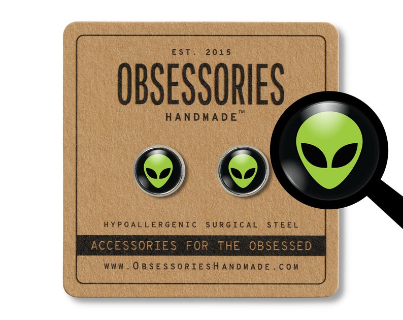 May include: A pair of hypoallergenic surgical steel earrings featuring a green alien head design. The earrings are packaged on a brown card with the text "OBSESSORIES HANDMADE" and "ACCESSORIES FOR THE OBSESSED".