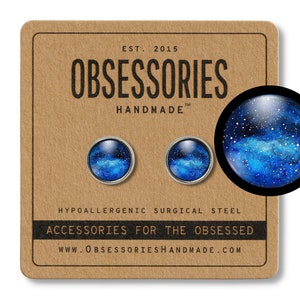 May include: A pair of stud earrings with a galaxy design. The earrings are made of hypoallergenic surgical steel and feature a blue and white watercolor galaxy design. The earrings are shown on a brown card with the text "OBSESSORIES HANDMADE" and "ACCESSORIES FOR THE OBSESSED".
