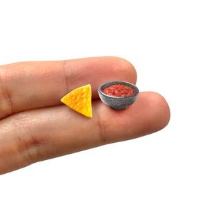 May include: Miniature food set featuring a single tortilla chip and a small bowl of salsa. The chip is a golden yellow color, and the salsa is a vibrant red. The bowl is a dark gray color.