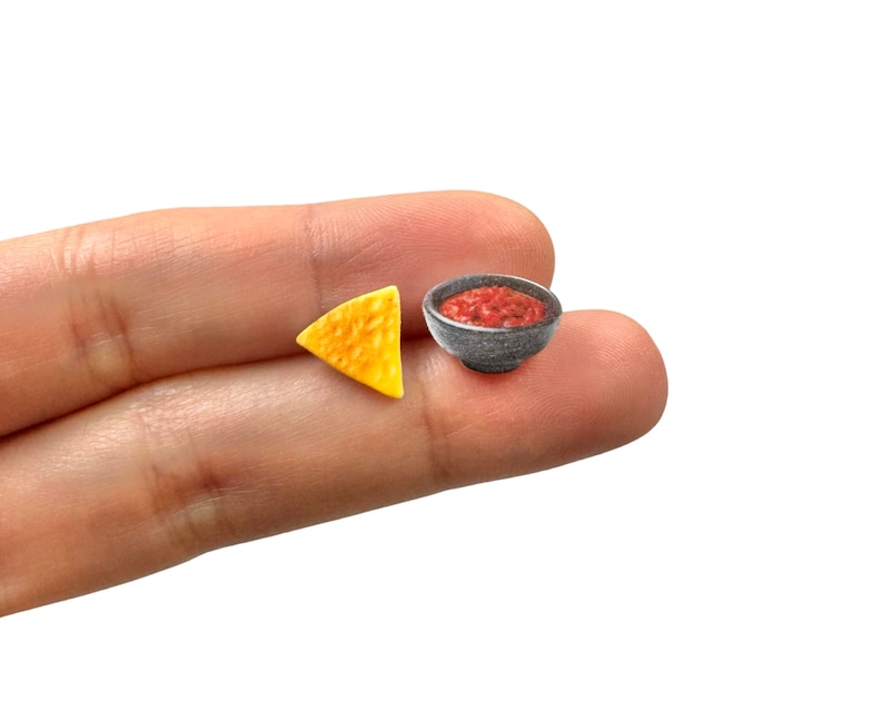 May include: Miniature food set featuring a single tortilla chip and a small bowl of salsa. The chip is a golden yellow color, and the salsa is a vibrant red. The bowl is a dark gray color.