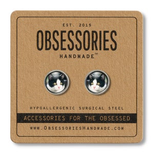 May include: A pair of hypoallergenic surgical steel stud earrings featuring a black and white cat face image. The earrings are packaged on a brown card with the text "OBSESSORIES HANDMADE" and "ACCESSORIES FOR THE OBSESSED".