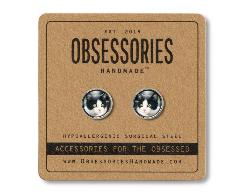 May include: A pair of hypoallergenic surgical steel stud earrings featuring a black and white cat face image. The earrings are packaged on a brown card with the text "OBSESSORIES HANDMADE" and "ACCESSORIES FOR THE OBSESSED".