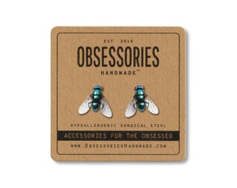 May include: A pair of fly-shaped earrings with teal and silver details, displayed on a brown card. The card reads "OBSESSORIES HANDMADE" and "ACCESSORIES FOR THE OBSESSED". The earrings are made of hypoallergenic surgical steel.