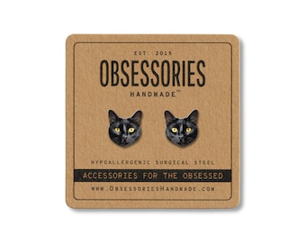 May include: A pair of black cat face stud earrings with yellow eyes on a brown card. The card reads "OBSESSORIES HANDMADE" and "ACCESSORIES FOR THE OBSESSED". The earrings are made of hypoallergenic surgical steel.