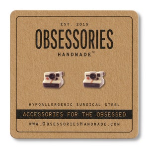 Puede incluir: Un par de pendientes con cámaras vintage en miniatura. Los pendientes están hechos de acero quirúrgico hipoalergénico y están etiquetados como "OBSESSORIES HANDMADE".