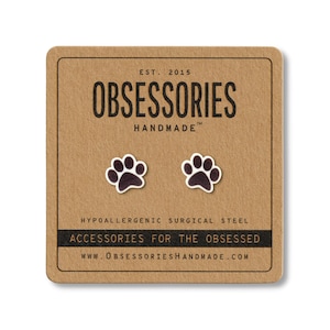 May include: A pair of black and white paw print stud earrings on a brown card. The card reads "OBSESSORIES HANDMADE" and "ACCESSORIES FOR THE OBSESSED". The earrings are made of hypoallergenic surgical steel.