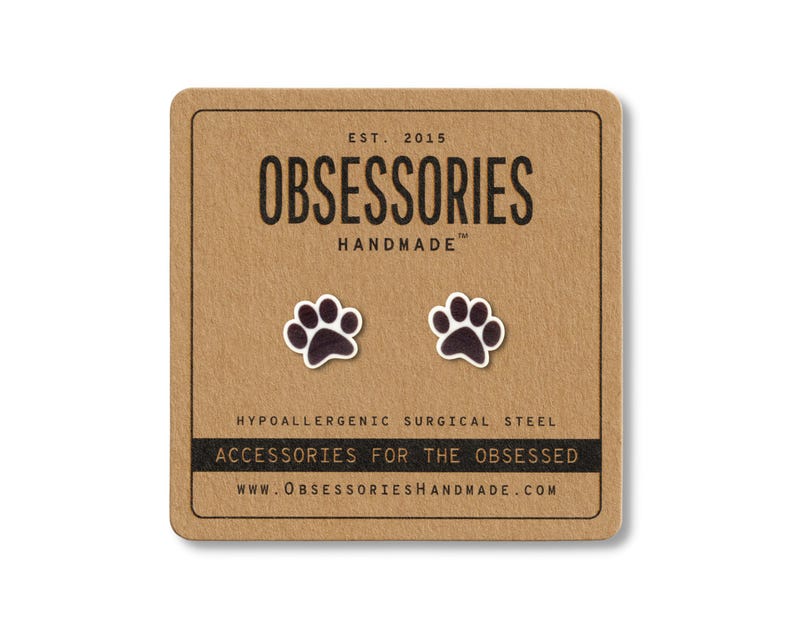 May include: A pair of black and white paw print stud earrings on a brown card. The card reads "OBSESSORIES HANDMADE" and "ACCESSORIES FOR THE OBSESSED". The earrings are made of hypoallergenic surgical steel.