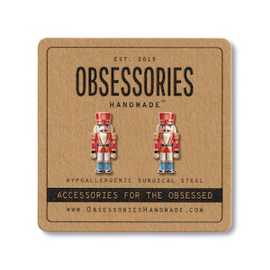 May include: A pair of nutcracker stud earrings made of surgical steel, displayed on a square brown cardboard card. The card features the text OBSESSORIES HANDMADE, EST. 2015, HYPOALLERGENIC SURGICAL STEEL, ACCESSORIES FOR THE OBSESSED, and the website URL.