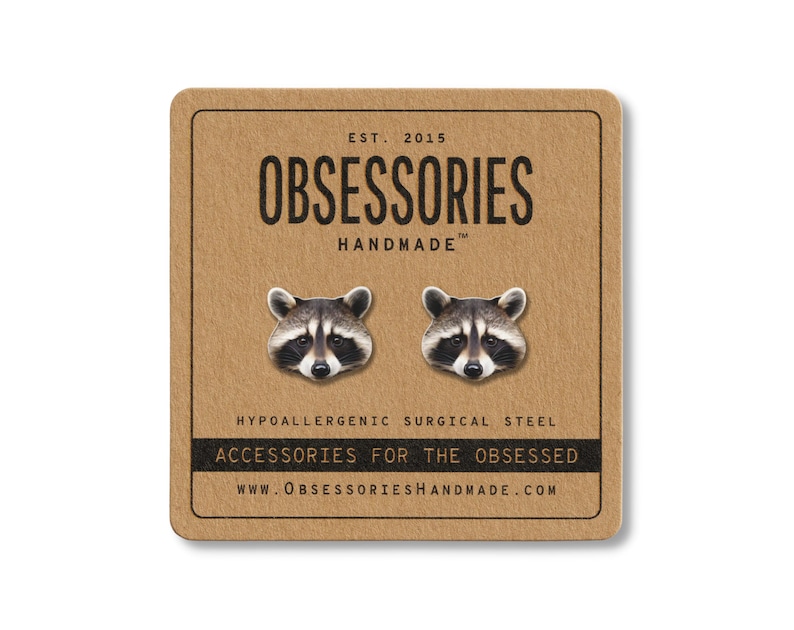 Puede incluir: Pendientes de bot&oacute;n con forma de cara de mapache sobre una tarjeta marr&oacute;n. La tarjeta dice "OBSESSORIES HANDMADE" y "ACCESSORIES FOR THE OBSESSED". Los pendientes son de acero quir&uacute;rgico hipoalerg&eacute;nico.