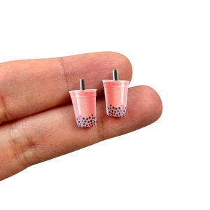 May include: Two small, pink bubble tea earrings with black tapioca pearls and black straws. The earrings are in the shape of a clear plastic cup. The earrings are held in a person's hand.