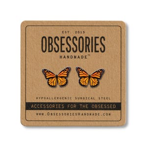 Puede incluir: Pendientes de mariposa con alas naranjas y negras, presentados en una tarjeta marrón. La tarjeta lleva el texto "OBSESSORIES HANDMADE" y "ACCESSORIES FOR THE OBSESSED". Los pendientes son de acero quirúrgico hipoalergénico.
