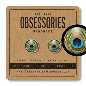 May include: A pair of hypoallergenic surgical steel earrings featuring a peacock feather design. The earrings are round and have a silver finish. The text "OBSESSORIES HANDMADE" is printed on a brown background.