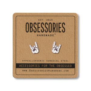 May include: Silver-coloured stud earrings in the shape of the rock and roll hand gesture, presented on a brown card. The card features the text "OBSESSORIES HANDMADE" and "ACCESSORIES FOR THE OBSESSED".