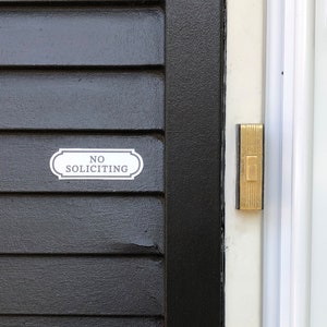 Puede incluir: Una puerta negra con un letrero blanco que dice "NO SOLICITING" y un bot&oacute;n de timbre dorado.