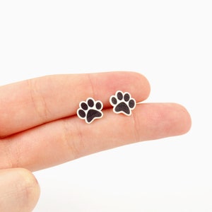 May include: Two small, paw-shaped earrings. Each earring features a black paw print design on a white background. The earrings are small and delicate, perfect for animal lovers.