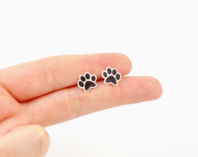May include: Two small, paw-shaped earrings. Each earring features a black paw print design on a white background. The earrings are small and delicate, perfect for animal lovers.