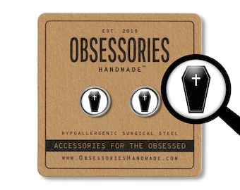 May include: A pair of silver cufflinks with black coffin designs and a white cross on each. The cufflinks are displayed on a brown card with the text "OBSESSORIES HANDMADE" and "ACCESSORIES FOR THE OBSESSED".