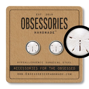 Pode incluir: Um par de brincos de aço cirúrgico hipoalergênico com um design de dólar de areia. Os brincos são feitos à mão e vêm em um cartão marrom com o texto "OBSESSORIES HANDMADE" e "ACCESSORIES FOR THE OBSESSED".