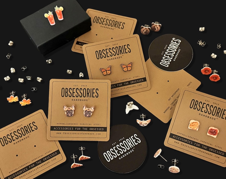 Puede incluir: Una colecci&oacute;n de pendientes de bot&oacute;n con diversos dise&ntilde;os, como mariposas, caras de perros y alimentos, presentados en tarjetas de marca. Las tarjetas dicen "OBSESSORIES HANDMADE". Tambi&eacute;n se ve una caja negra y cierres de pendientes.