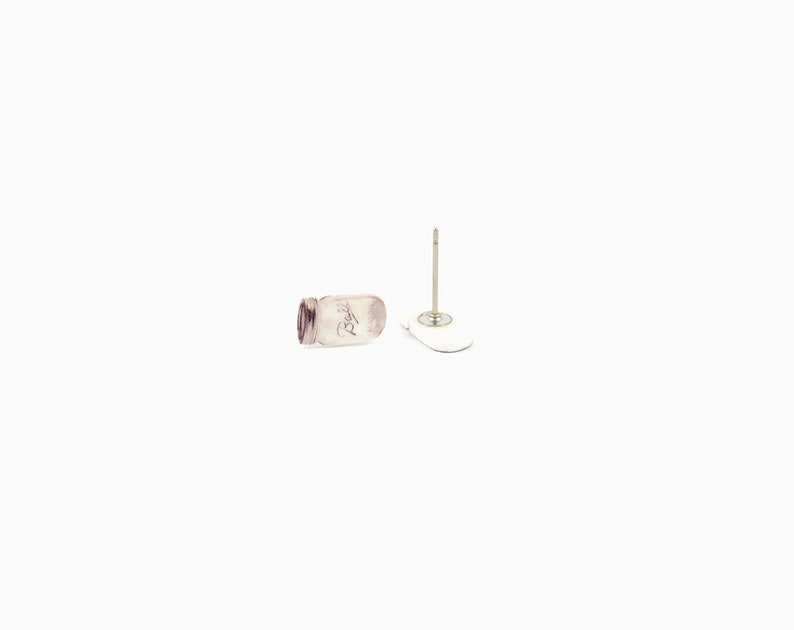 May include: A pair of stud earrings featuring a small, clear mason jar design. One earring is lying on its side, while the other is upright with a post and a white backing. The earrings are set against a white background.