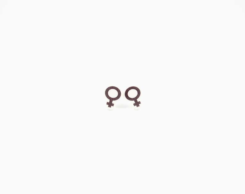 May include: Two brown glittery earrings in the shape of the female gender symbol, set against a white background. The earrings are small and detailed, with a textured appearance. The design is simple and elegant.