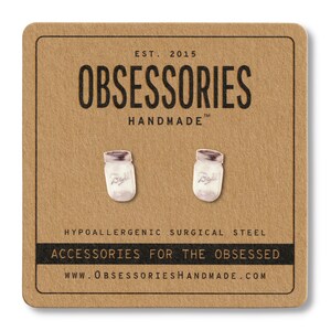 May include: A pair of silver-colored jar earrings on a brown card. The earrings are shaped like mason jars with a white interior and a silver lid. The card has the text "OBSESSORIES HANDMADE" and "ACCESSORIES FOR THE OBSESSED".