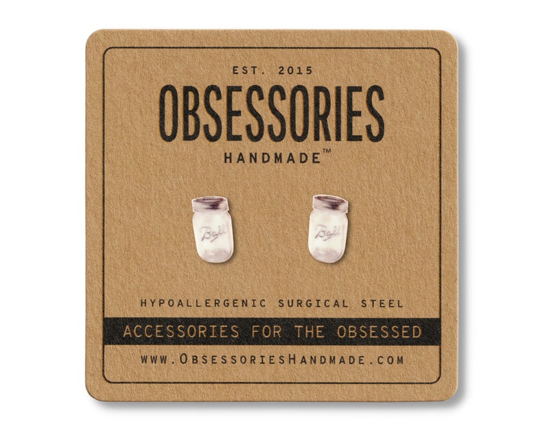 May include: A pair of silver-colored jar earrings on a brown card. The earrings are shaped like mason jars with a white interior and a silver lid. The card has the text "OBSESSORIES HANDMADE" and "ACCESSORIES FOR THE OBSESSED".