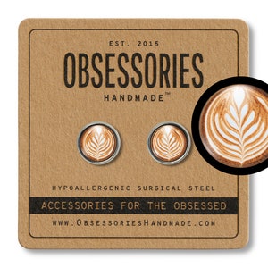 Puede incluir: Un par de pendientes de acero quirúrgico hipoalergénico con un diseño de arte latte. Los pendientes están hechos a mano y presentan un diseño de arte latte marrón y blanco con un patrón de hojas. Los pendientes están empaquetados en una tarjeta marrón con el texto "OBSESSORIES HANDMADE" y "ACCESSORIES FOR THE OBSESSED".
