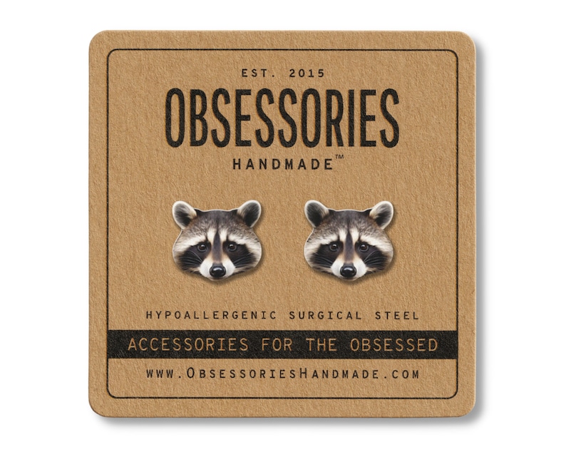 Puede incluir: Pendientes de bot&oacute;n con cabeza de mapache sobre una tarjeta marr&oacute;n. Los pendientes son plateados con caras de mapache detalladas. La tarjeta tiene el texto "OBSESSORIES HANDMADE" y "ACCESSORIES FOR THE OBSESSED".