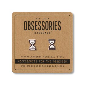 May include: A pair of silver-colored hourglass stud earrings on a brown card. The card reads "OBSESSORIES HANDMADE" and "ACCESSORIES FOR THE OBSESSED". The earrings are made of hypoallergenic surgical steel.