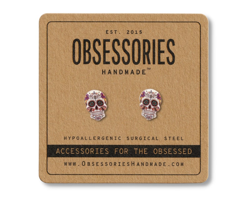 May include: A pair of skull-shaped earrings with colorful floral designs, displayed on a brown card. The card features the text "OBSESSORIES HANDMADE" and "ACCESSORIES FOR THE OBSESSED". The earrings are made of hypoallergenic surgical steel.