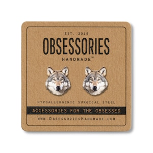 May include: A pair of wolf head stud earrings on a brown card. The earrings are silver and gray, with detailed wolf faces. The card reads "OBSESSORIES HANDMADE" and "ACCESSORIES FOR THE OBSESSED."