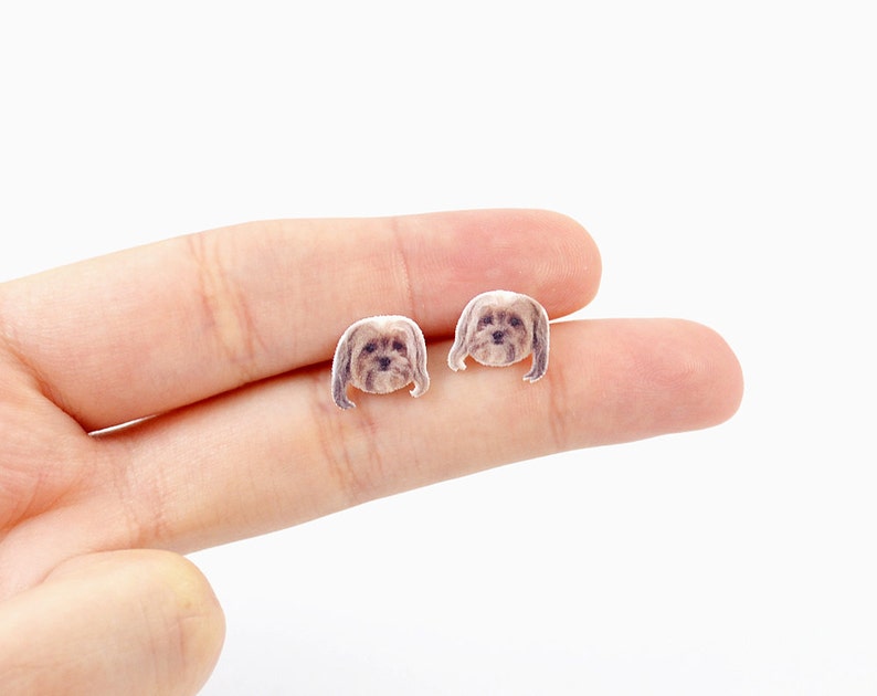 May include: A pair of small, dog-shaped stud earrings. The earrings feature a detailed illustration of a dog's face with brown and tan fur. The earrings are held in a person's hand against a white background.
