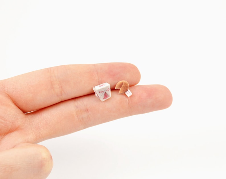 May include: Miniature Chinese takeout box with a red and white design, next to a fortune cookie with a white paper fortune. The box has the word "Enjoy" printed on it. The items are small and appear to be dollhouse accessories.