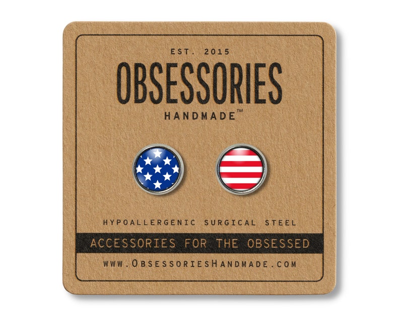 Pode incluir: Um par de pequenos brincos redondos prateados com um design de bandeira americana azul e branca em um brinco e um design de listras vermelhas e brancas no outro brinco. Os brincos est&atilde;o em um cart&atilde;o marrom com o texto "OBSESSORIES HANDMADE" e "ACCESSORIES FOR THE OBSESSED".