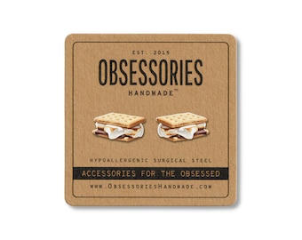 May include: A square, brown card with the text "OBSESSORIES HANDMADE" and two s'mores illustrations. The card also includes the text "ACCESSORIES FOR THE OBSESSED" and "WWW.OBSESSORIESHANDMADE.COM".