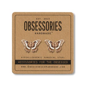 Może przedstawiać: Kolczyki w kształcie motyli na brązowej karcie. Kolczyki są brązowe, białe i beżowe. Na karcie widnieje napis "OBSESSORIES HANDMADE" i "ACCESSORIES FOR THE OBSESSED". Kolczyki wykonane są z hipoalergicznej stali chirurgicznej.