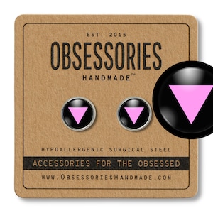 Puede incluir: Un par de pendientes de botón negros y plateados con un símbolo de triángulo rosa. Los pendientes son hipoalergénicos y están hechos de acero quirúrgico. El embalaje es marrón con el texto "OBSESSORIES HANDMADE" y "ACCESSORIES FOR THE OBSESSED".