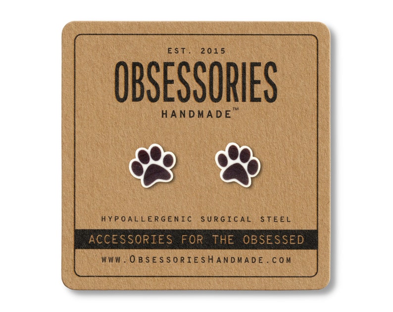 May include: A pair of paw print stud earrings on a brown card. The earrings are dark brown with white outlines. The card has the text "OBSESSORIES HANDMADE" and "ACCESSORIES FOR THE OBSESSED".