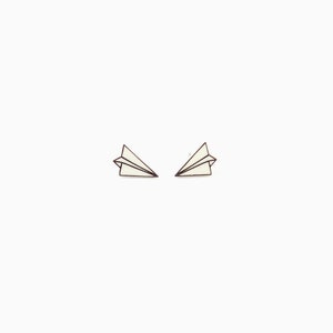 May include: Two paper airplane earrings. Each earring is a small, white paper airplane with black outline details. The earrings are set against a plain white background.