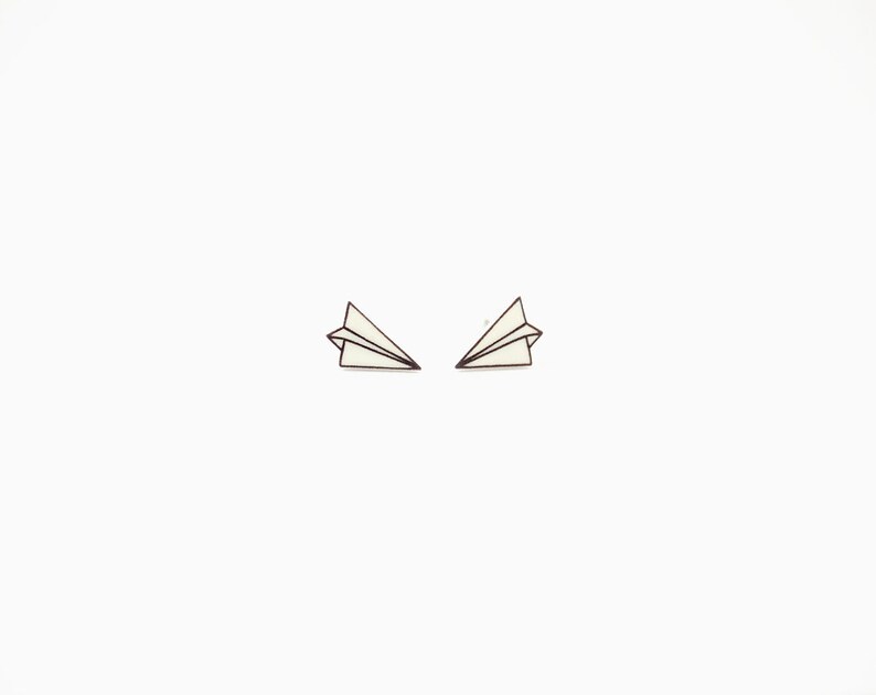 May include: Two paper airplane earrings. Each earring is a small, white paper airplane with black outline details. The earrings are set against a plain white background.