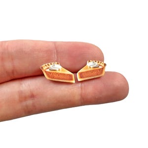 May include: Two miniature pie slice earrings. Each earring features a slice of pie with a golden-brown crust, a creamy white dollop of whipped cream, and a filling of orange-brown color. The earrings are small and detailed.