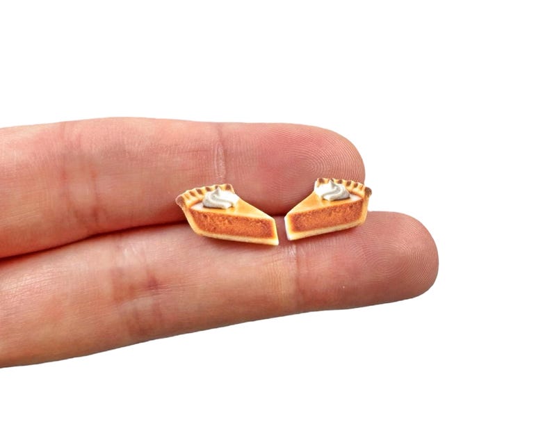 May include: Two miniature pie slice earrings. Each earring features a slice of pie with a golden-brown crust, a creamy white dollop of whipped cream, and a filling of orange-brown color. The earrings are small and detailed.