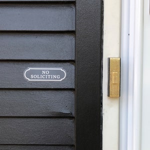 Puede incluir: Una puerta negra con un letrero blanco que dice "No Soliciting" y un bot&oacute;n de timbre dorado.