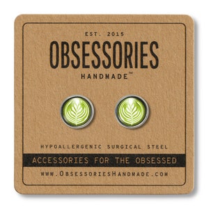 May include: A pair of silver stud earrings with a green latte art design. The earrings are hypoallergenic and made of surgical steel. The packaging features the text "OBSESSORIES HANDMADE" and "ACCESSORIES FOR THE OBSESSED".