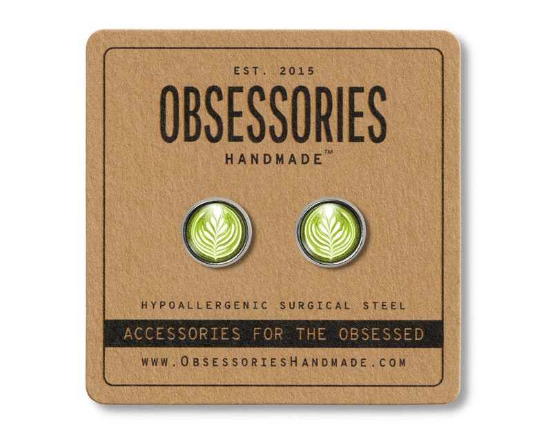 May include: A pair of silver stud earrings with a green latte art design. The earrings are hypoallergenic and made of surgical steel. The packaging features the text "OBSESSORIES HANDMADE" and "ACCESSORIES FOR THE OBSESSED".