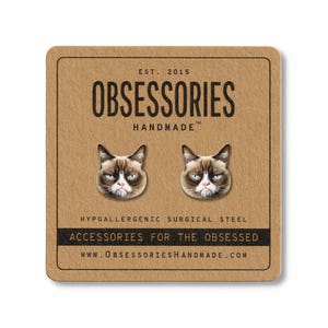May include: A pair of stud earrings featuring the face of a grumpy cat. The earrings are on a brown square card with the text "OBSESSORIES HANDMADE" and "ACCESSORIES FOR THE OBSESSED". The card also includes the website address.