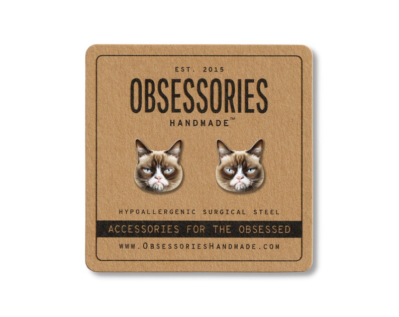 May include: A pair of stud earrings featuring the face of a grumpy cat. The earrings are on a brown square card with the text "OBSESSORIES HANDMADE" and "ACCESSORIES FOR THE OBSESSED". The card also includes the website address.