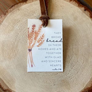 以下が含まれることがあります： 茶色のベルベットのリボンが付いた長方形のタグ。水彩画の小麦のイラストと「They broke bread in their homes and ate together with glad and sincere hearts acts 2:46」というテキストが特徴です。